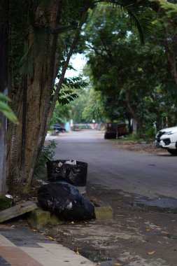 Çöp dolu büyük siyah bir plastik torba ağaçlı sessiz bir otobanın yanında yer almaktadır.