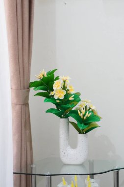 Odanın köşesine temiz ve estetik bir dekorasyon olarak yerleştirilmiş eşsiz bir çömleğe yerleştirilmiş yapay frangipani çiçekleri.