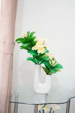 Odanın köşesine temiz ve estetik bir dekorasyon olarak yerleştirilmiş eşsiz bir çömleğe yerleştirilmiş yapay frangipani çiçekleri.