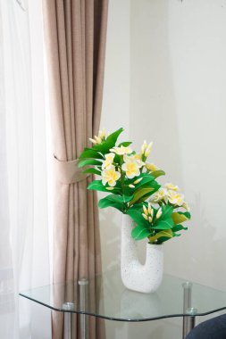 Odanın köşesine temiz ve estetik bir dekorasyon olarak yerleştirilmiş eşsiz bir çömleğe yerleştirilmiş yapay frangipani çiçekleri.