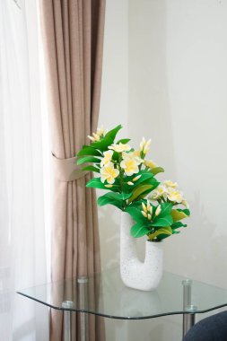 Odanın köşesine temiz ve estetik bir dekorasyon olarak yerleştirilmiş eşsiz bir çömleğe yerleştirilmiş yapay frangipani çiçekleri.