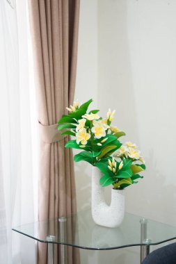 Odanın köşesine temiz ve estetik bir dekorasyon olarak yerleştirilmiş eşsiz bir çömleğe yerleştirilmiş yapay frangipani çiçekleri.
