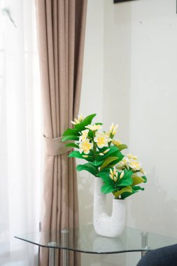 Odanın köşesine temiz ve estetik bir dekorasyon olarak yerleştirilmiş eşsiz bir çömleğe yerleştirilmiş yapay frangipani çiçekleri.