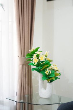 Odanın köşesine temiz ve estetik bir dekorasyon olarak yerleştirilmiş eşsiz bir çömleğe yerleştirilmiş yapay frangipani çiçekleri.