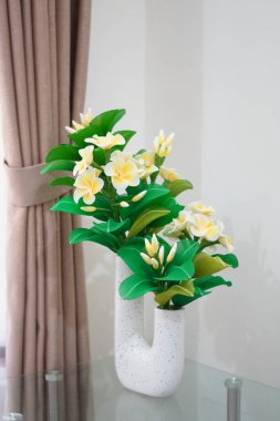 Odanın köşesine temiz ve estetik bir dekorasyon olarak yerleştirilmiş eşsiz bir çömleğe yerleştirilmiş yapay frangipani çiçekleri.