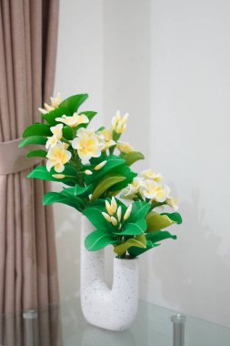Odanın köşesine temiz ve estetik bir dekorasyon olarak yerleştirilmiş eşsiz bir çömleğe yerleştirilmiş yapay frangipani çiçekleri.