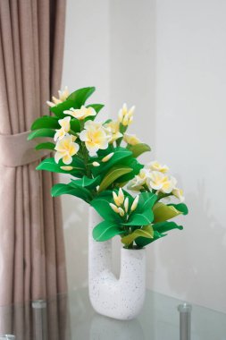 Odanın köşesine temiz ve estetik bir dekorasyon olarak yerleştirilmiş eşsiz bir çömleğe yerleştirilmiş yapay frangipani çiçekleri.