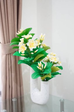Odanın köşesine temiz ve estetik bir dekorasyon olarak yerleştirilmiş eşsiz bir çömleğe yerleştirilmiş yapay frangipani çiçekleri.