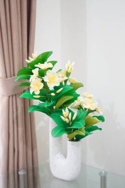 Odanın köşesine temiz ve estetik bir dekorasyon olarak yerleştirilmiş eşsiz bir çömleğe yerleştirilmiş yapay frangipani çiçekleri.
