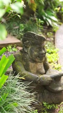 Balili bir adam şeklinde oyulmuş taş bir heykel geleneksel bir Balili müzik aleti taşıyor bahçe süsü olarak.