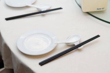 Beyaz tabaklar, çorba kaşıkları ve yemek çubukları bir restoranda beyaz masa örtüsüyle yuvarlak bir masada.