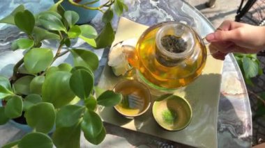 Çay takımı, cam çaydanlık ve fincanlar. Bira. Yavaş çekim çayı. kompozisyon. Yeşil otelin yeşil çayı yasemin çaydanlığı iki fincana doluyor verandada, çiçek saksılarının üzerinde. Güzellik Yasemin