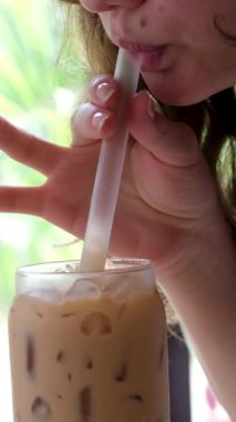 Kadın elleri açık hava restoranında metal pipetle kokteyl karıştırıyor. Güneşli tropikal kafede çikolatalı milkshake içen genç bir kadın. Masadaki kız büyük bardakta buzlu kahve içiyor.. 