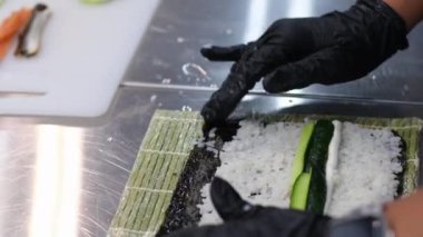 Profesyonel bir suşi şefinin elleri Norveç yapraklarının üzerine pirinç seriyor beyaz mutfak masasının üzerinde bambu bir hasırın üzerine uzanmış bir sıra taze sebze, biber ve marul yapraklarıyla birlikte..