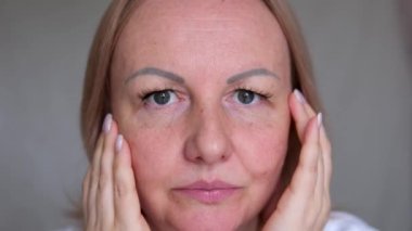 Yaşlı kadın nemlendirici krem kullanıyor. Son sınıf dişisi el ve parmaklarıyla yaşlanma önleyici losyon sürüyor, yanağına dokunuyor, anti-yaş yüz bakımı uyguluyor. Yüzünde kırışıklıklar, sivilceler ve sivilceler.