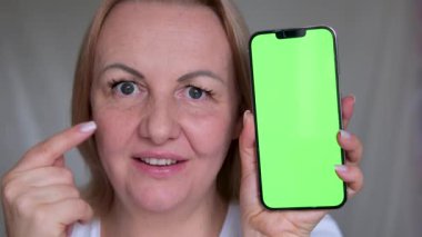 Krom tuşlu akıllı telefonlu uygulamayı tarayan bir kız evde online sipariş veriyor. Mutfaktaki yeşil ekran cep telefonuna dokunan kadın. Kadın parmakları kapalı alandaki cep telefonunu işaret ediyor.. 