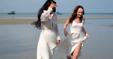Sahilde yürüyen iki kız arkadaş LGBT beyaz elbiseler giyip dans ediyorlar. Asya 'lı güzel genç kadınlar, deniz nehrinin sığ sularında dans ediyorlar. Yüksek kalite