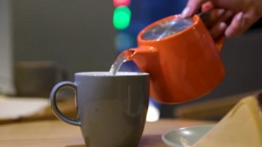 Ev hanımı çaydanlıktan, içinde çay poşeti olan şeffaf fincana sıcak su döker. Kadın, mutfakta öğle yemeği molası için aromatik amfibik çay demler. Yüksek kalite 4k görüntü
