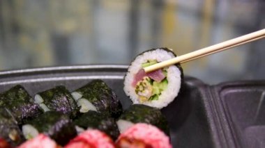 Çubuklar suşi kutusuna bir suşi koydu. Plastik bir kutuda yuvarlanıyor. Çeşitli lezzetli suşi setleri bir teslimat konteynerinde. Konsept Asya yemekleri, sağlıklı yiyecekler, paket yemek..