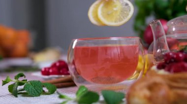 Portakallı, limonlu ve limonlu çayı şeffaf çaydanlıktan bardağa doldurmak. Arka planda bal ve krep ile çay demlemek. Yavaş çekim çayı. Kırmızı amberli iki dilim limon.