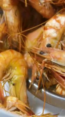 Kırmızı karideslerin dikey videoda kaynatıldığı tavanın yanındaki çiğ büyük kaplan karidesi kasesi. Küçük mutfakta elektrikli fırında lezzetli protein zengini kabuklular pişiriyorlar. Yüksek kalite