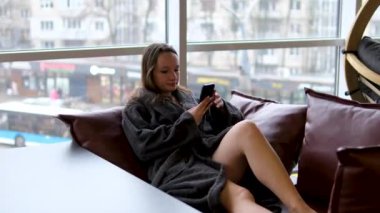 Chambers ev kıyafetleri duştan çık pencerenin yanındaki kanepeye otur mutlu, gamsız, neşeli, beyaz kadın kız. cep telefonu kullanımı akıllı telefon rahatlatma sosyal medya sohbet