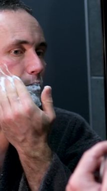 Yaşlı adam aynadaki yansımasına bakıyor ve tıraş oluyor. Günlük sabah prosedürünü kabul et. Sabahlık giyen adam aynanın yanında spa tedavisi görüyor köpük güzellik salonunu tıraş ediyor ev banyosunda kendine bakıyor.