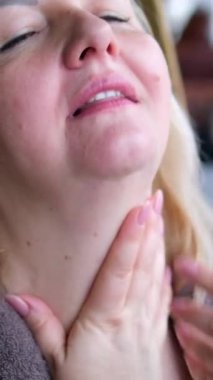 Orta yaşlı bir kadın boynundaki kremi kaplıcaya benzetiyor. Esmer saçı görünüyor ve elleri cilt bakımına karışıyor. Boyun masajı.