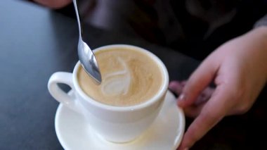 Tahta masada beyaz bardakta kaşıkla karıştırılmış koyu köpüklü sıcak kahve kapuçinosu, yakın çekim. Sütlü kapuçino, latte. Yüksek kalite 4k görüntü