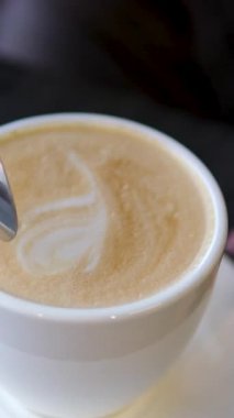 Tahta masada beyaz bardakta kaşıkla karıştırılmış koyu köpüklü sıcak kahve kapuçinosu, yakın çekim. Sütlü kapuçino, latte. Yüksek kalite 4k görüntü