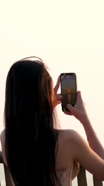 Günbatımını akıllı telefonlu bir kızla fotoğraflayan kadın okyanus manzarası üzerinde saçlarını uçuran yumuşak ışıklı bir mercek manzarası manzara manzarası tatil macerasının tadını çıkaran bir kadın. .
