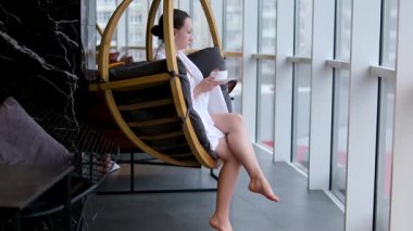 Beyaz bornozlu bir kadın büyük panoramik pencerenin yanında çarşaflı bir şekilde oturuyor. Yanında da bir fincan çıplak ayakla sepetin içinde. Yüksek kalite 4k görüntü