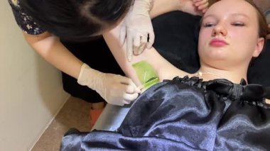 Güzellik uzmanı müşterilerine tahtadan bir spatula ile sıcak balmumu uygular. Balmumuyla deplasman. Balmumu Koltuk Altı Tüyleri Depilasyonu. Epilasyon Ağrısı Olan Kadın