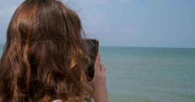 Gezgin kadın deniz kenarında oturur ve cep telefonu kullanır. Yüksek kaliteli FullHD görüntüler