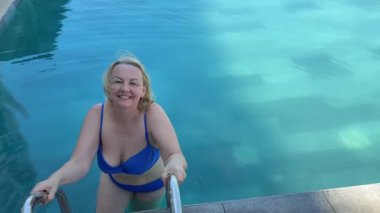 Yetişkin, tombul bir kadın havuza rahatlamak için girer mavi mayolu, sarı saçlı, 45-55 yaşlarında güzel bir gülümseme. Yüksek kalite 4k görüntü
