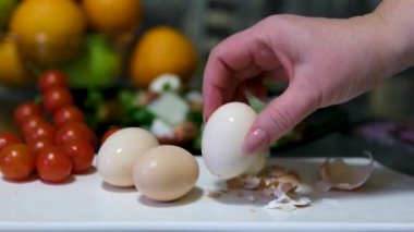 Açık tenli, haşlanmış yumurtayı tabağın üzerine soyan, içinde yumurta kabuğu parçaları olan, beyaz gömlek ve önlük giyen, kahverengi saçlı, başka bir tabağın kenarından aşağı ve aşağıya doğru akan bulanık bir kadının yakın çekimi.. 