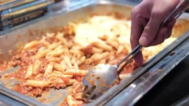 Açık büfe makarna servis ediliyor. İnsanlar uzun bir masanın etrafında iki sıra halinde, içinde alfredo, marinara sosu, pirinç, sebze ve tavuk olan makarna servis ediyorlar..