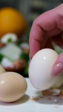 Açık tenli, haşlanmış yumurtayı tabağın üzerine soyan, içinde yumurta kabuğu parçaları olan, beyaz gömlek ve önlük giyen, kahverengi saçlı, başka bir tabağın kenarından aşağı ve aşağıya doğru akan bulanık bir kadının yakın çekimi.. 