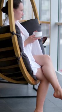 Beyaz bornozlu bir kadın büyük panoramik pencerenin yanında çarşaflı bir şekilde oturuyor. Yanında da bir fincan çıplak ayakla sepetin içinde. Yüksek kalite 4k görüntü