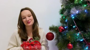 Kameraya hediyesini gösteren güzel ve çekici esmer kadın. Beyaz, güzel bir kadın Noel ağacında oturuyor. Gülümseyen kız hediyesi Noel
