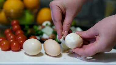Kadın cam tabakta yumurta kabuğunu kırıyor sonra mutfak masasında yumurta kabuğunu kabuğundan soyuyor önlük giyip patates ve yemek pişiriyor.