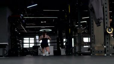 Jimnastik salonu öne doğru hareket ediyor, ayakları ve bacakları esnetiyor, parlak spor salonunda ileri atlıyor, beyaz atlet ve siyah şort giyiyor, arka planda geniş pencereler ve ekipman rafları var, ısınma hareketleri sergiliyor..