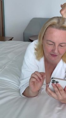 Beyaz bornozlu bir kadın, güzel dekore edilmiş bir yatak odasındaki bir yatakta otururken akıllı telefon kullanıyor. Rahat bir otel odasındaki bir yatağın üzerinde telefon kullanarak bornozlu kadını rahatlatıyor.