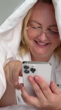 Beyaz bornozlu bir kadın, güzel dekore edilmiş bir yatak odasındaki bir yatakta otururken akıllı telefon kullanıyor. Rahat bir otel odasındaki bir yatağın üzerinde telefon kullanarak bornozlu kadını rahatlatıyor.