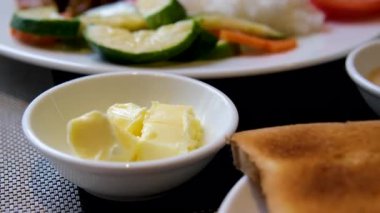 Bir insan taze sebzeler, turşular ve otlarla çevrili iyi aydınlatılmış bir kahvaltı masasında bir dilim rustik ekmeğin üzerine tereyağı sürer. Doğal ışıklandırma, yakın çekim açısı. Yüksek kalite 4k görüntü