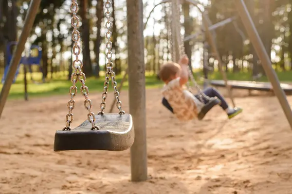Küçük bir çocuk parkta yerde oturuyor, ön planda boş bir salıncak.