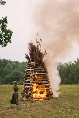 Geleneksel kostümlü bir kadın elinde bir saman yığınıyla yanan ateşin yanında duruyor.