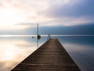Bir ahşap iskele, yatın güneşin doğuşuna ve sisin içindeki dağlara karşı durduğu göle kadar gider.