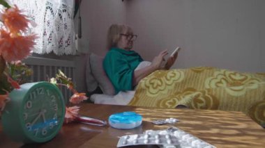 Yaşlı, gözlüklü hasta bir kadın yatakta yatarken telefona bakar. Evde tedavi edilen hasta bir emeklinin portresi. Tedavi kavramı.