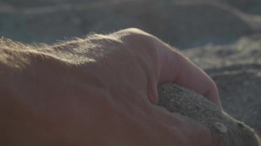 Bir adamın eli kumsalda, güneş ışığında parmaklarının arasından kum döker. İnce kum taneleri güneşin altındaki sıcak altın kumun üzerine yavaşça düşer gün batımında parlar. Deniz kıyısında dinlen, deniz kenarında dinlen.